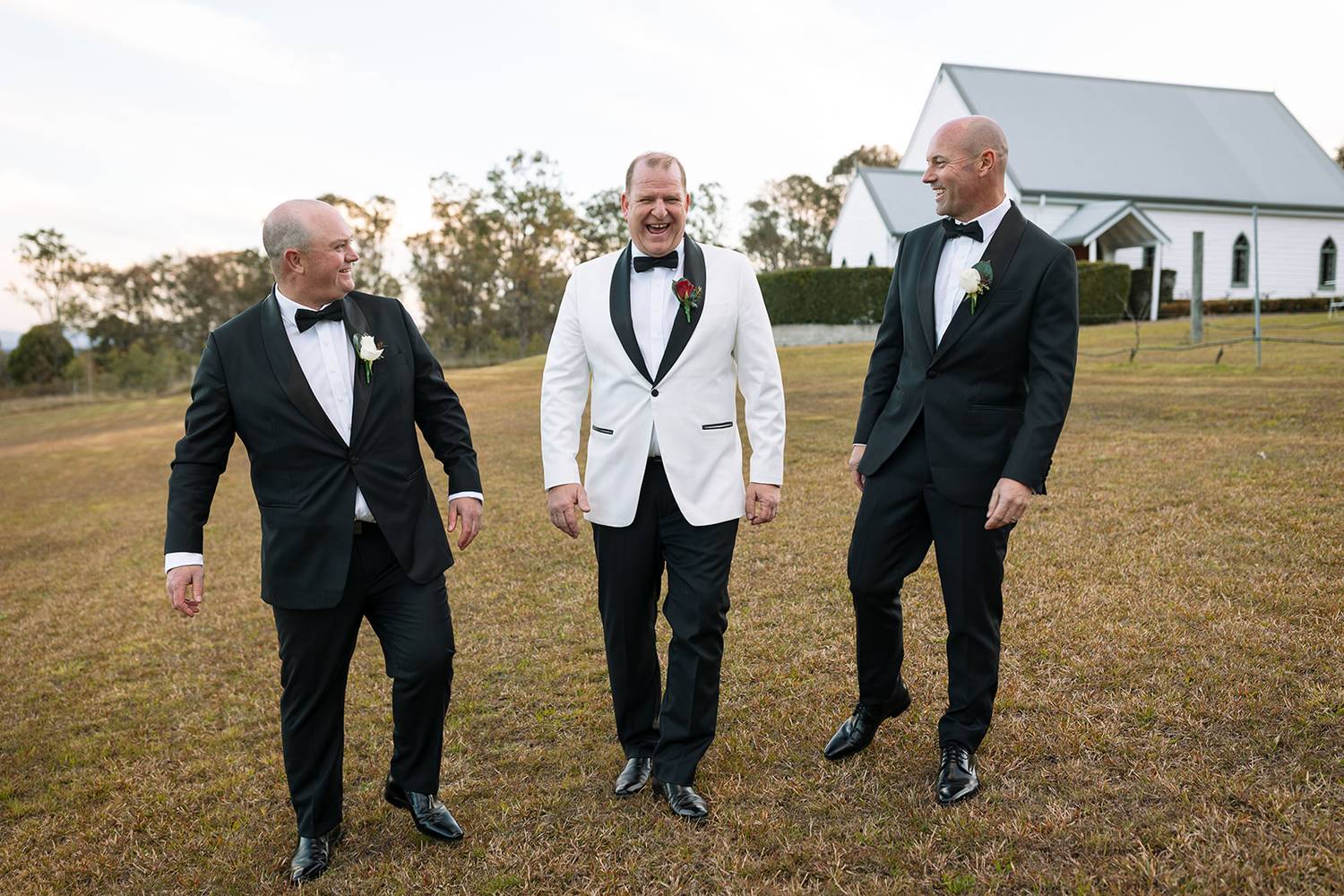 Sven in an ivory tuxedo with a black satin shawl lapel laughing with two groomsmen in black tuxedos beside Lovedale Wedding Chapel in the Hunter Valley